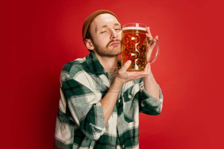 Foto de Retrato de un joven con camisa a cuadros casual posando con cerveza lager aislada sobre fondo rojo. Amante de las bebidas. Concepto de emociones, gusto, estilo de vida, expresión facial, Oktoberfest - Imagen libre de derechos