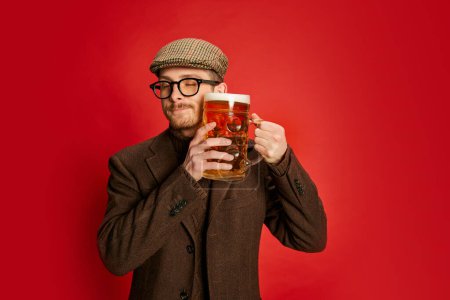 Téléchargez les photos : Homme élégant en vêtements classiques posant avec un verre de bière mousseuse lager isolé sur fond rouge. Vendredi soir rafraîchissement. Concept d'émotions, goût, style de vie, expression faciale, Oktoberfest - en image libre de droit