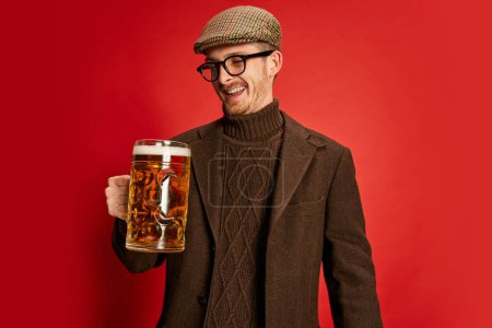 Téléchargez les photos : Portrait d'homme élégant dans des vêtements classiques posant avec un verre de bière mousseuse lager isolé sur fond rouge. Goût délicieux. Concept d'émotions, goût, style de vie, expression faciale, Oktoberfest - en image libre de droit