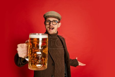 Téléchargez les photos : Portrait d'homme élégant dans des vêtements classiques posant avec un verre de bière mousseuse lager isolé sur fond rouge. À la tienne. Concept d'émotions, goût, style de vie, expression faciale, Oktoberfest - en image libre de droit