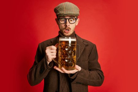 Foto de Retrato de un hombre con estilo en ropa clásica posando con un vaso de cerveza espumante lager aislado sobre fondo rojo. Cara conmocionada. Concepto de emociones, gusto, estilo de vida, expresión facial, Oktoberfest - Imagen libre de derechos
