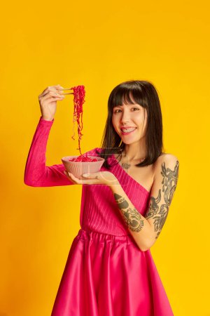 Photo for Asian food lover. Young beautiful girl in bright pink dress eating pink noodles isolated on vivid yellow background. Concept of youth, beauty, fashion, lifestyle, emotions, facial expression. Ad - Royalty Free Image