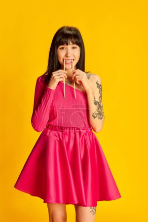 Téléchargez les photos : Portrait de jeune fille drôle en robe rose vif posant avec des baguettes dans la bouche isolé sur fond jaune vif. Concept de jeunesse, beauté, mode, mode de vie, émotions, expression faciale. Publicité - en image libre de droit