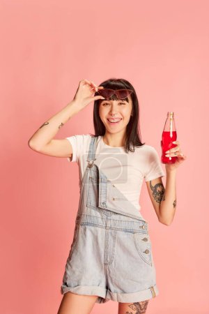 Photo for Young stylish girl posing in denim overalls, holding soda and cheerfully laughing isolated on pink background. Summer vacation. Youth, beauty, fashion, lifestyle, emotions, facial expression concept - Royalty Free Image