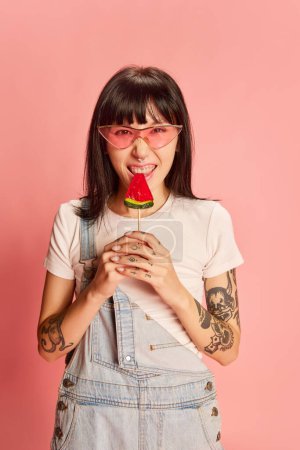 Photo for Young stylish girl in trendy sunglasses, with tattoos, posing in denim overalls, eating lollipop isolated on pink background. Concept of youth, beauty, fashion, lifestyle, emotions, facial expression - Royalty Free Image
