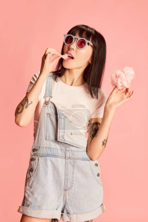 Photo for Young stylish girl posing in denim overalls, eating cotton candy isolated on pink background. Summer fun. Concept of youth, beauty, fashion, lifestyle, emotions, facial expression. Ad - Royalty Free Image