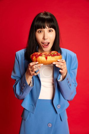 Téléchargez les photos : Portrait de jeune fille en costume bleu élégant posant, mangeant hot-dog épicé isolé sur fond rouge. Pause déjeuner. Concept de jeunesse, beauté, mode, mode de vie, émotions, expression faciale. Publicité - en image libre de droit