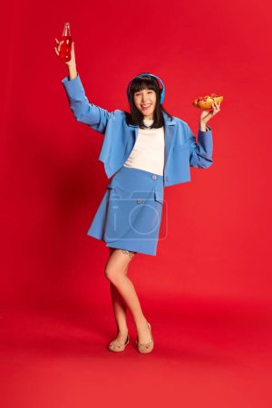 Téléchargez les photos : Joyeux jeune fille en costume bleu élégant posant dans des écouteurs avec hotdog isolé sur fond rouge. Concept de jeunesse, beauté, mode, mode de vie, émotions, expression faciale. Publicité - en image libre de droit