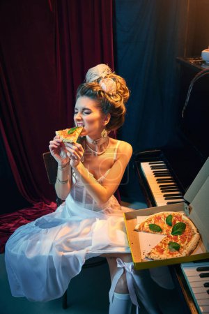 Foto de Amante de la pizza. Retrato de una joven hermosa niña en imagen de persona medieval en elegante vestido blanco sentado al piano con comida italiana. Comparación de épocas, belleza, historia, arte, creatividad. - Imagen libre de derechos