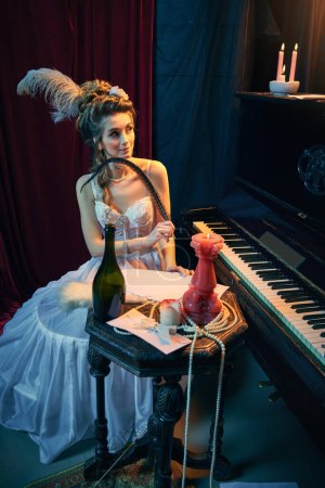 Foto de Retrato de una joven hermosa niña en imagen de persona medieval en elegante vestido blanco sentado al piano y escribiendo novela. Comparación de épocas, belleza, historia, arte, creatividad. - Imagen libre de derechos