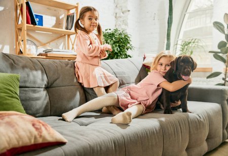 Foto de Dos niñas encantadoras, niños jugando, abrazando perro de raza pura, labrador marrón en casa. Divirtiéndose. Concepto de familia, infancia, mascotas, cuidado, amistad, emociones y tiempo libre - Imagen libre de derechos
