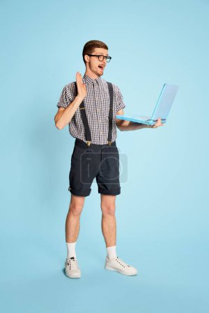 Photo for Young man in checkered shirt, shorts and suspenders having video call on laptop isolated over blue background. Internet meeting. Concept of emotions, business, education, occupation, facial expression - Royalty Free Image