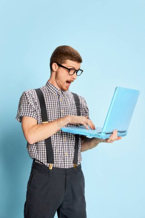 Téléchargez les photos : Jeune homme en chemise à carreaux et bretelles tapant émotionnellement sur ordinateur portable isolé sur fond bleu. Développeur. Concept d'émotions, d'affaires, d'éducation, d'occupation, d'expression faciale - en image libre de droit