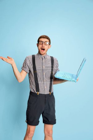 Téléchargez les photos : Jeune homme émotif en chemise à carreaux, short et bretelles posant ordinateur portable isolé sur fond bleu. Achats en ligne. Concept d'émotions, d'affaires, d'éducation, d'occupation, d'expression faciale - en image libre de droit