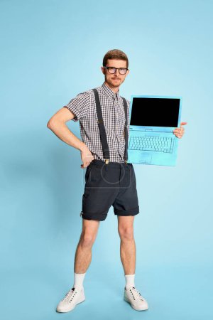 Téléchargez les photos : Jeune homme en chemise à carreaux, short et bretelles posant avec ordinateur portable isolé sur fond bleu. Grandes ventes. Concept d'émotions, d'affaires, d'éducation, d'occupation, d'expression faciale - en image libre de droit