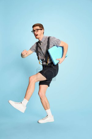 Photo for Portrait of young man in checkered shirt, shorts and suspenders with laptop isolated over blue background. Positivity. Concept of emotions, business, education, occupation, facial expression - Royalty Free Image