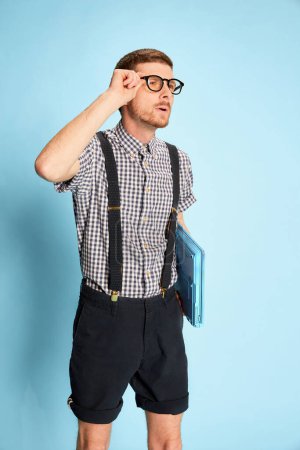 Photo for Portrait of young man, student in checkered shirt, shorts and suspenders with laptop isolated over blue background. Concept of emotions, business, education, occupation, facial expression - Royalty Free Image