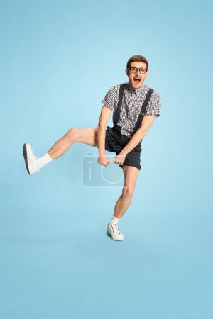 Téléchargez les photos : Portrait de jeune homme en chemise à carreaux, short et bretelles sautant joyeusement isolé sur fond bleu. Succès. Concept d'émotions, d'affaires, d'éducation, d'occupation, d'expression faciale - en image libre de droit