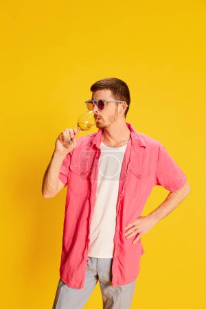 Photo for Portrait of young man in pink shirt posing isolated over vivid yellow background. Wine degustation. Concept of youth, lifestyle, alcohol, casual fashion, emotions, facial expression. Ad - Royalty Free Image
