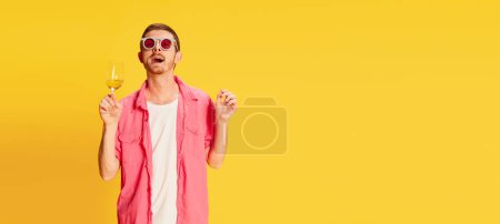 Foto de Hombre joven con camisa rosa, gafas de sol divertidas posando con copa de vino aislada sobre fondo amarillo. Relajado. Concepto de juventud, estilo de vida, alcohol, moda, emociones, expresión facial. Copiar espacio para anuncio - Imagen libre de derechos