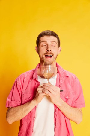 Photo for Portrait of young man in pink shirt posing with wine glass isolated over vivid yellow background. Smelling good. Concept of youth, lifestyle, alcohol, casual fashion, emotions, facial expression. Ad - Royalty Free Image