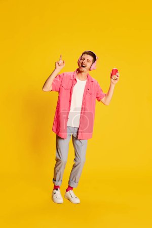 Photo for Summer party. Young man in pink shirt posing, listening to music in headphones isolated on vivid yellow background. Concept of youth, lifestyle, music, casual fashion, emotions, facial expression. Ad - Royalty Free Image