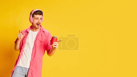 Téléchargez les photos : Jeune homme émotif en chemise rose posant dans des écouteurs avec soda isolé sur fond jaune vif. Flyer. Concept de jeunesse, mode de vie, musique, mode, émotions, expression faciale. Espace de copie pour la publicité - en image libre de droit