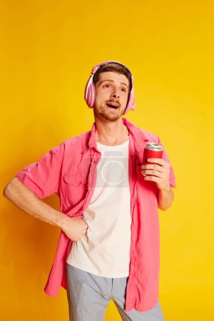 Téléchargez les photos : Portrait de jeune homme en chemise rose posant isolé sur fond jaune vif. Musique et soda. Heureux. Concept de jeunesse, style de vie, fête, mode décontractée, émotions, expression faciale. Publicité - en image libre de droit