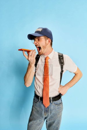 Foto de Retrato de un joven, estudiante posando en ropa casual, gritando por teléfono con ira aislada sobre fondo azul. Concepto de juventud, estilo de vida, educación, emociones, expresión facial. Anuncio - Imagen libre de derechos