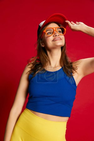 Photo for Portrait of young beautiful girl in sportive, blue and yellow clothes posing over red background. Positive energy. Concept of youth, beauty, sport lifestyle, emotions, facial expression. Ad - Royalty Free Image