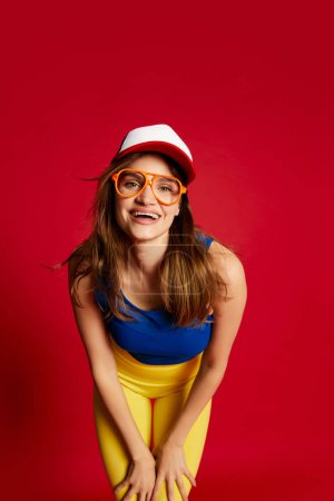 Photo for Portrait of young beautiful girl in sportive clothes, cap and glasses posing over red background. Happy. Concept of youth, beauty, sport lifestyle, emotions, fitness, facial expression. Ad - Royalty Free Image