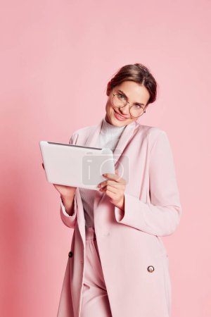 Photo for Portrait of beautiful young girl in suit and glasses posing, typing, working on tablet over pink background. Manager. Concept of youth, beauty, fashion, lifestyle, emotions, facial expression. Ad - Royalty Free Image