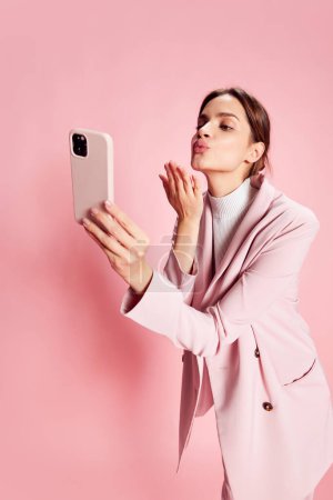 Photo for Portrait of young beautiful girl in a suit, having video call on phone over pink background. Sending kisses. Concept of youth, beauty, fashion, lifestyle, emotions, facial expression. Ad - Royalty Free Image