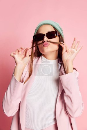 Foto de Retrato de una joven hermosa niña en traje, sombrero y gafas de sol posando sobre fondo rosa. Buen humor. Concepto de juventud, belleza, moda, estilo de vida, emociones, expresión facial. Anuncio - Imagen libre de derechos