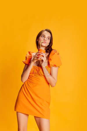 Téléchargez les photos : Portrait de belle fille brune en robe mignonne posant, buvant de la coke sur fond orange. Délicieux. Concept de jeunesse, beauté, mode, mode de vie, émotions, expression faciale. Publicité - en image libre de droit