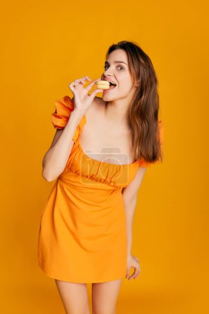 Foto de Retrato de una hermosa chica morena en lindo vestido posando, comiendo macaron sobre fondo naranja. Dulce diente. Concepto de juventud, belleza, moda, estilo de vida, emociones, expresión facial. Anuncio - Imagen libre de derechos