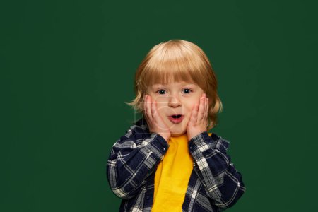 Photo for Portrait of little boy, child om yellow T-shirt and checkered shirt posing over green studio background. Happy shock. Concept of childhood, emotions, lifestyle, fashion, happiness. Copy space for ad - Royalty Free Image