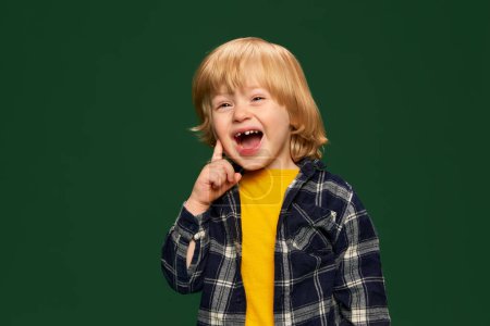 Téléchargez les photos : Portrait de petit garçon heureux, enfant posant sur fond de studio vert. Joyeux rire. Des blagues drôles. Concept d'enfance, émotions, mode de vie, mode, bonheur. Espace de copie pour la publicité - en image libre de droit
