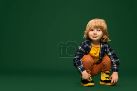 Photo for Portrait of cute little boy, child posing, sitting on squats over green studio background. Calm baby. Concept of childhood, emotions, lifestyle, fashion, happiness. Copy space for ad - Royalty Free Image