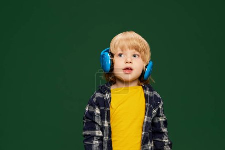 Photo for Portrait of little boy, child posing in headphones, listening to fairy tales over green studio background. Concept of childhood, emotions, lifestyle, fashion, happiness. Copy space for ad - Royalty Free Image