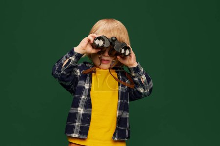 Photo for Portrait of little boy, child posing, looking in binoculars over green studio background. Adventures, game. Concept of childhood, emotions, lifestyle, fashion, happiness. Copy space for ad - Royalty Free Image