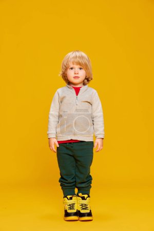 Téléchargez les photos : Portrait de petit garçon, enfant posant dans des vêtements chauds confortables sur fond de studio jaune. Du calme. Concept d'enfance, émotions, mode de vie, mode, bonheur. Espace de copie pour la publicité - en image libre de droit