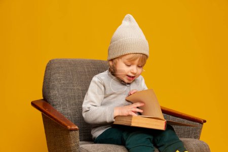 Photo for Portrait of little boy, child posing, sitting on chair and reading book over yellow studio background. Fairy tales. Concept of childhood, emotions, lifestyle, fashion, happiness. Copy space for ad - Royalty Free Image