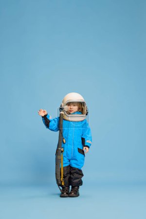 Téléchargez les photos : Portrait de petit garçon, enfant posant en costume d'astronaute sur fond bleu studio. Jouer à des jeux. Concept d'enfance, émotions, mode de vie, mode, bonheur. Espace de copie pour la publicité - en image libre de droit