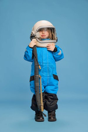 Téléchargez les photos : Portrait de petit garçon, enfant posant en costume d'astronaute sur fond bleu studio. Enfant émotionnel. Concept d'enfance, émotions, mode de vie, mode, bonheur. Espace de copie pour la publicité - en image libre de droit