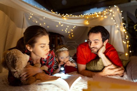 Photo for Lovely family, mother, father and daughter lying inside self-made hut, tent in room in the evening and reading book, fairy tale. Concept of fantasy, childhood, family, leisure time, love, care, home. - Royalty Free Image
