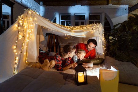 Photo for Lovely family, mother, father, daughter lying inside self-made hut, tent in room in the evening with Christmas lights and reading tales. Concept of fantasy, childhood, leisure time, love, care, home. - Royalty Free Image
