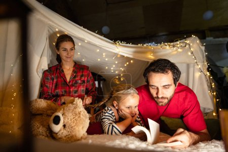 Photo for Loving family. Mother, father and daughter lying inside self-made hut, tent with lights, toys and pillows and reading book. Fairy tales. Concept of fantasy, childhood, leisure time, love, care, home. - Royalty Free Image