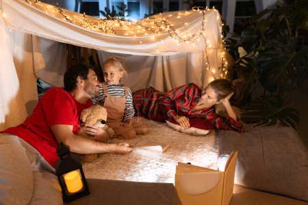 Photo for Pajama party. Mother, father and daughter lying inside self-made hut, tent in room in the evening and talking. Concept of fantasy, childhood, family, leisure time, love, care, home. - Royalty Free Image