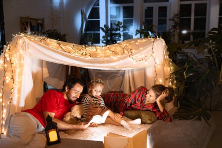Photo for Mother, father and daughter lying on pillows inside self-made hut, tent in room in the evening with Christmas lights. Concept of fantasy, childhood, family, leisure time, love, care, home. - Royalty Free Image